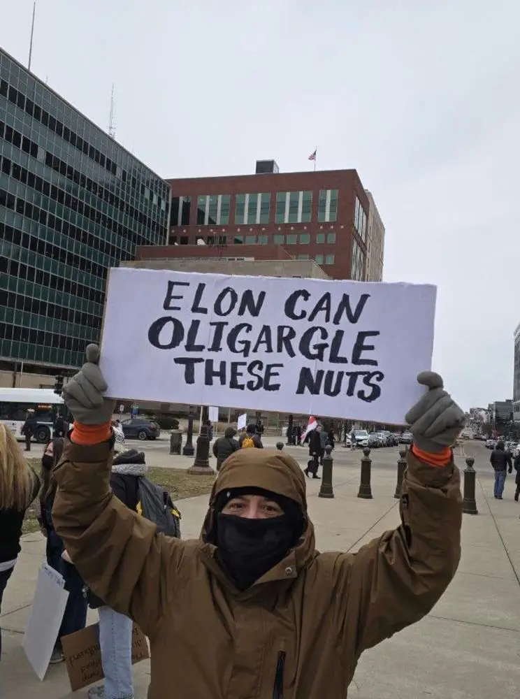 A person wearing a brown hooded jacket and black face mask holds up a handmade white sign with black handwritten text that…