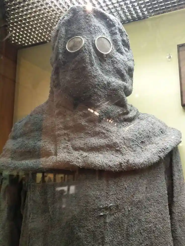 A close-up of a museum exhibit showing a full-body, gray knitted garment with a hood and two round, clear goggles embedded…