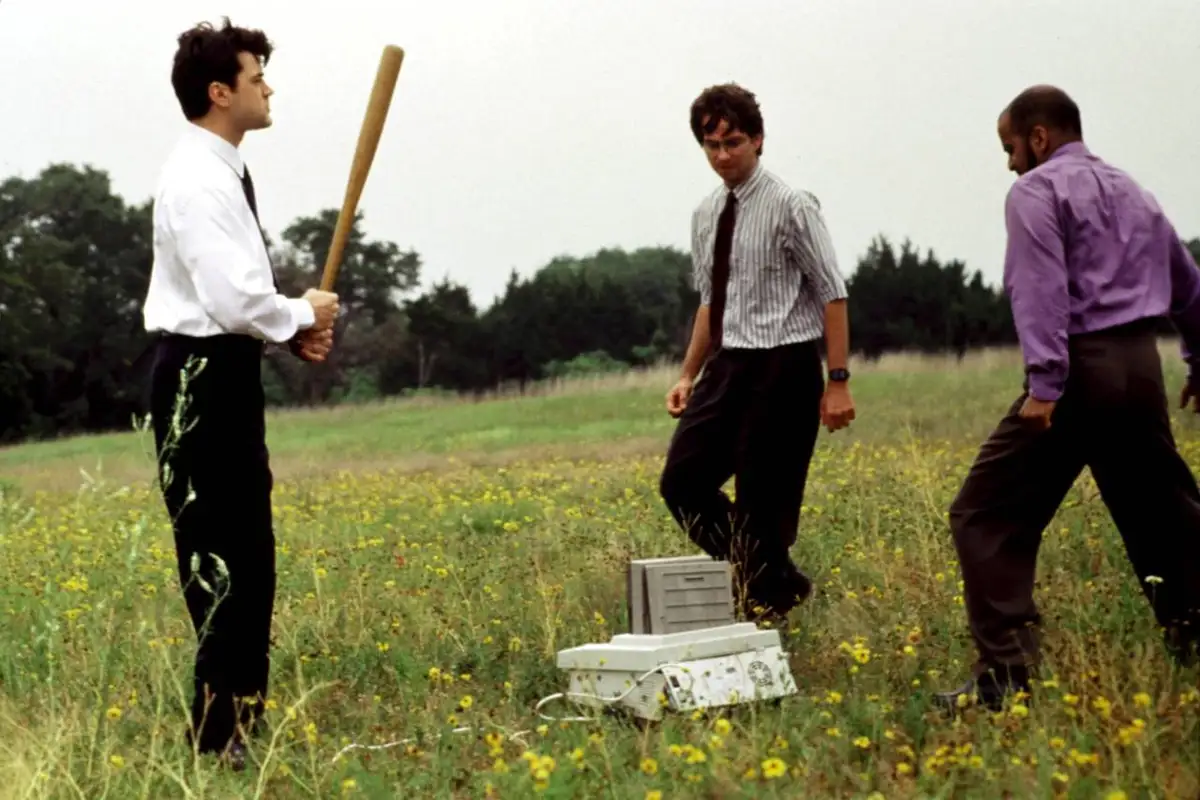 Three men in business attire stand in a grassy field with yellow wildflowers. One man on the left holds a wooden baseball…
