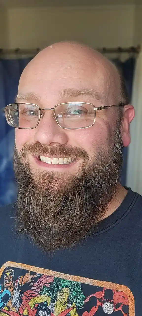 A close-up photo of a smiling bald man with a full brown beard and glasses, wearing a dark t-shirt with a colorful comic…