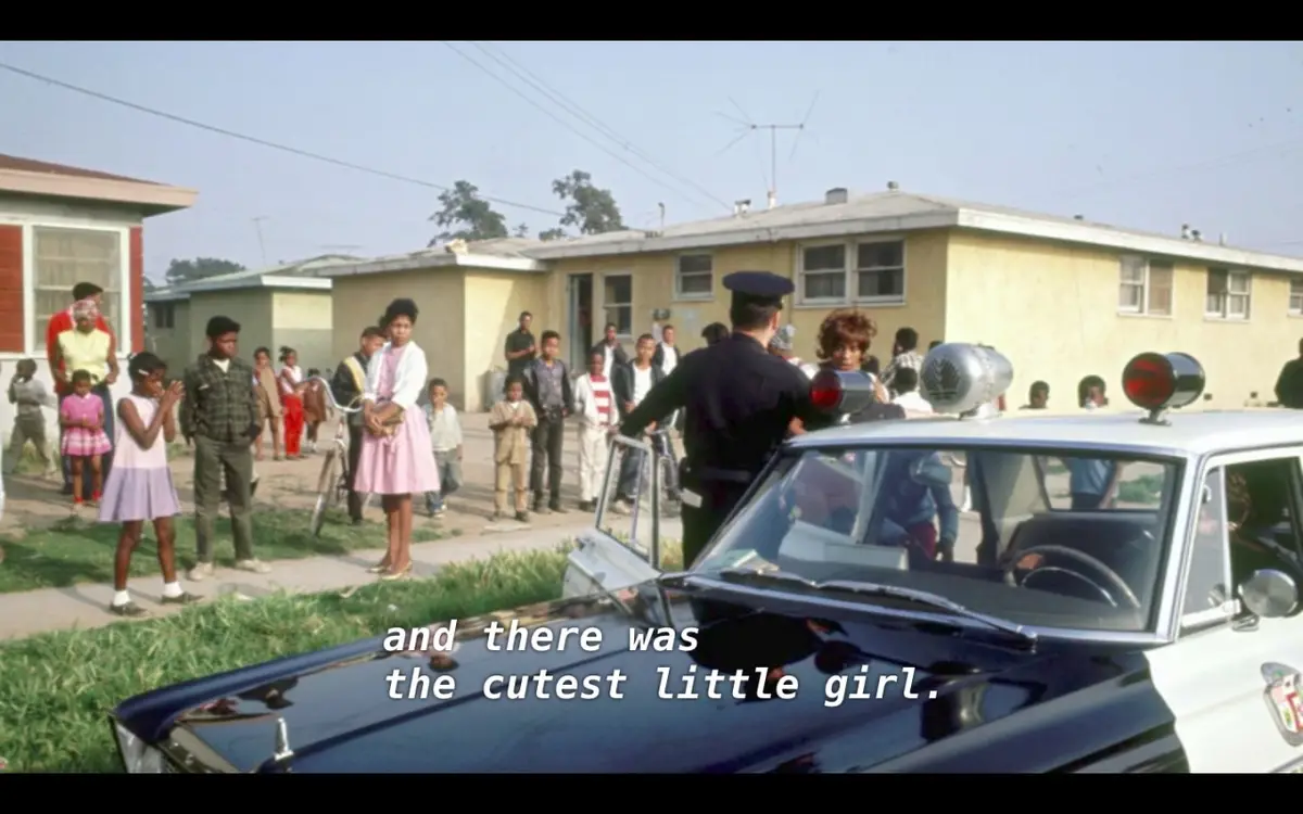 A vintage photo shows a police officer standing beside a classic police car with its door open, facing a crowd of onlookers…