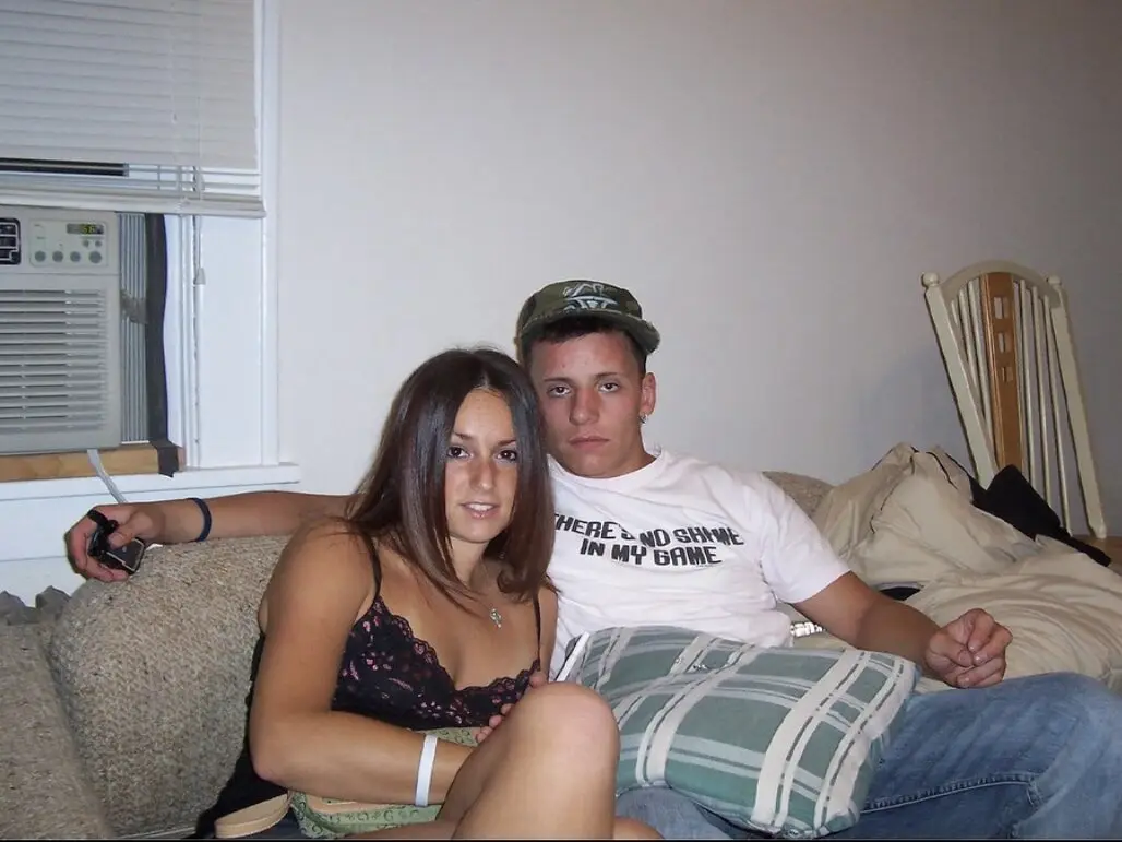 A young man and woman sit on a couch in a casual living room setting. The man, wearing a camouflage cap and a white t-shirt…