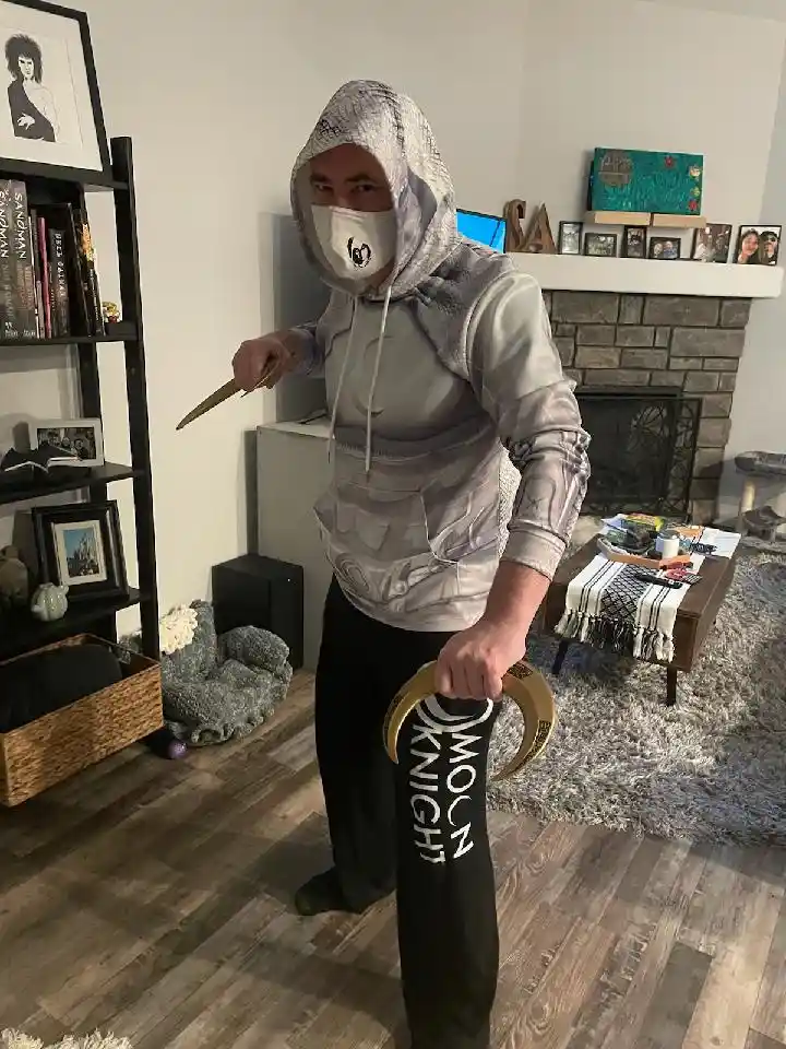 A person in a grey hooded sweatshirt with a printed knight armor design and a white face mask stands in a living room…
