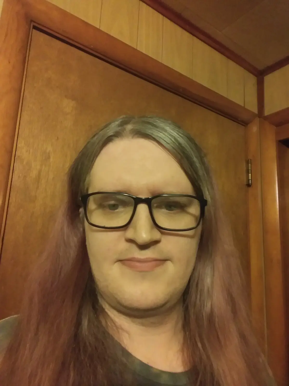 A person with long, multi-toned hair (blonde, pink, and brown) and black-rimmed glasses stares directly at the camera with a…
