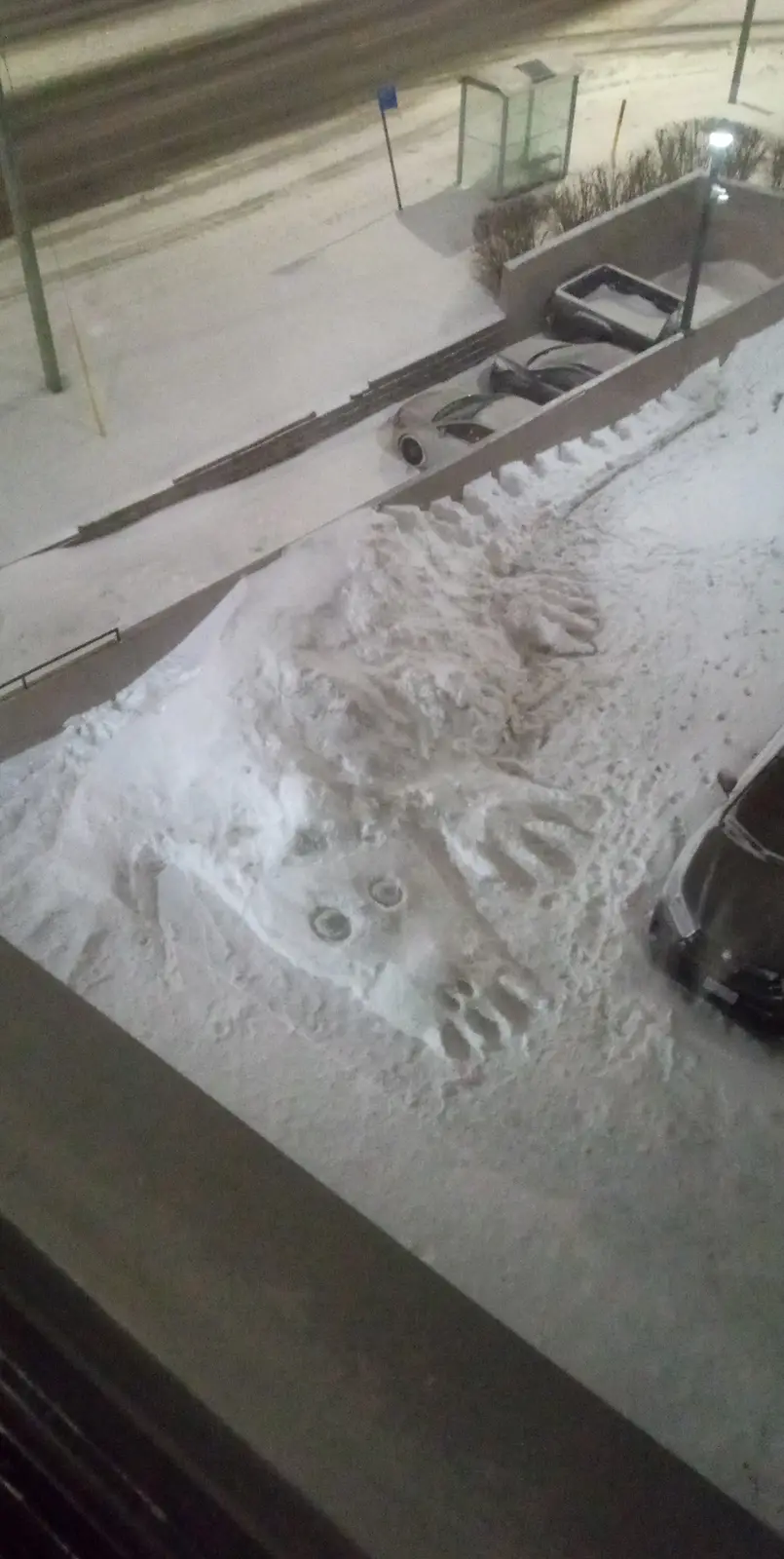 An overhead nighttime photo shows a large pile of snow on a sidewalk with distinct handprints and two circular indentations…