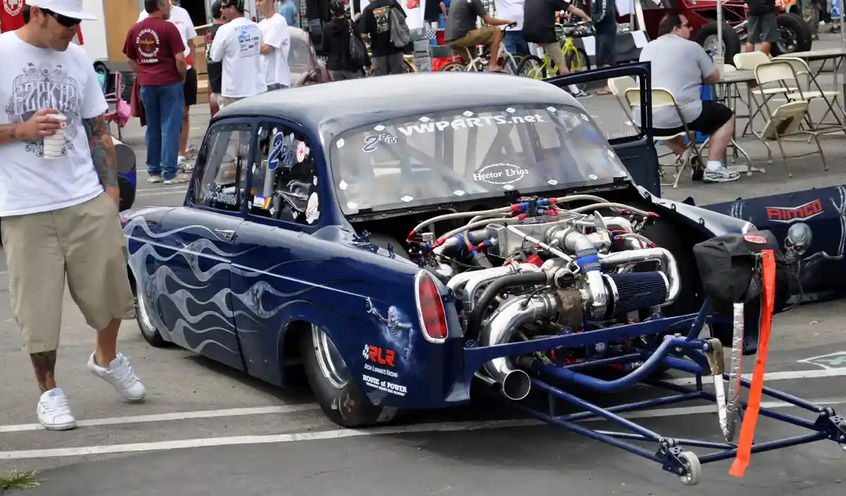 A heavily modified blue hot rod with a massive exposed engine and flame decals is parked at an outdoor car show. A man in a…