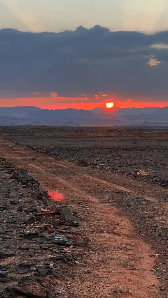 A wide desert landscape at sunset with a bright orange sun setting behind distant mountains, casting a warm glow over a dirt…