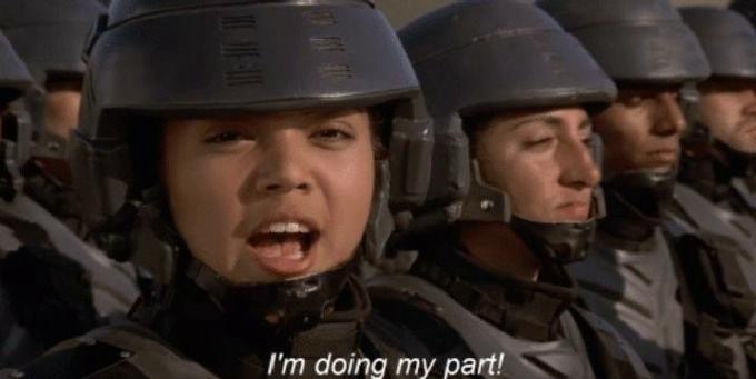 A close-up of a female soldier in a dark helmet and tactical gear, mouth open as if shouting, with other soldiers in similar…