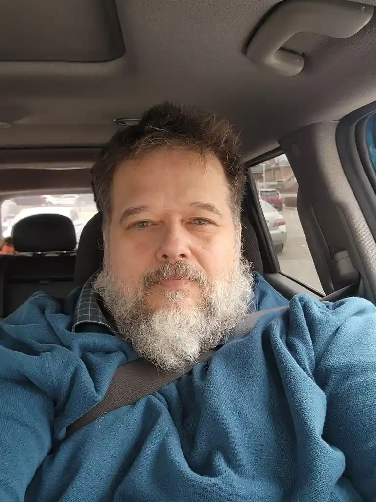 A man with a full, graying beard and mustache sits in the driver's seat of a car, wearing a blue fleece hoodie over a plaid…