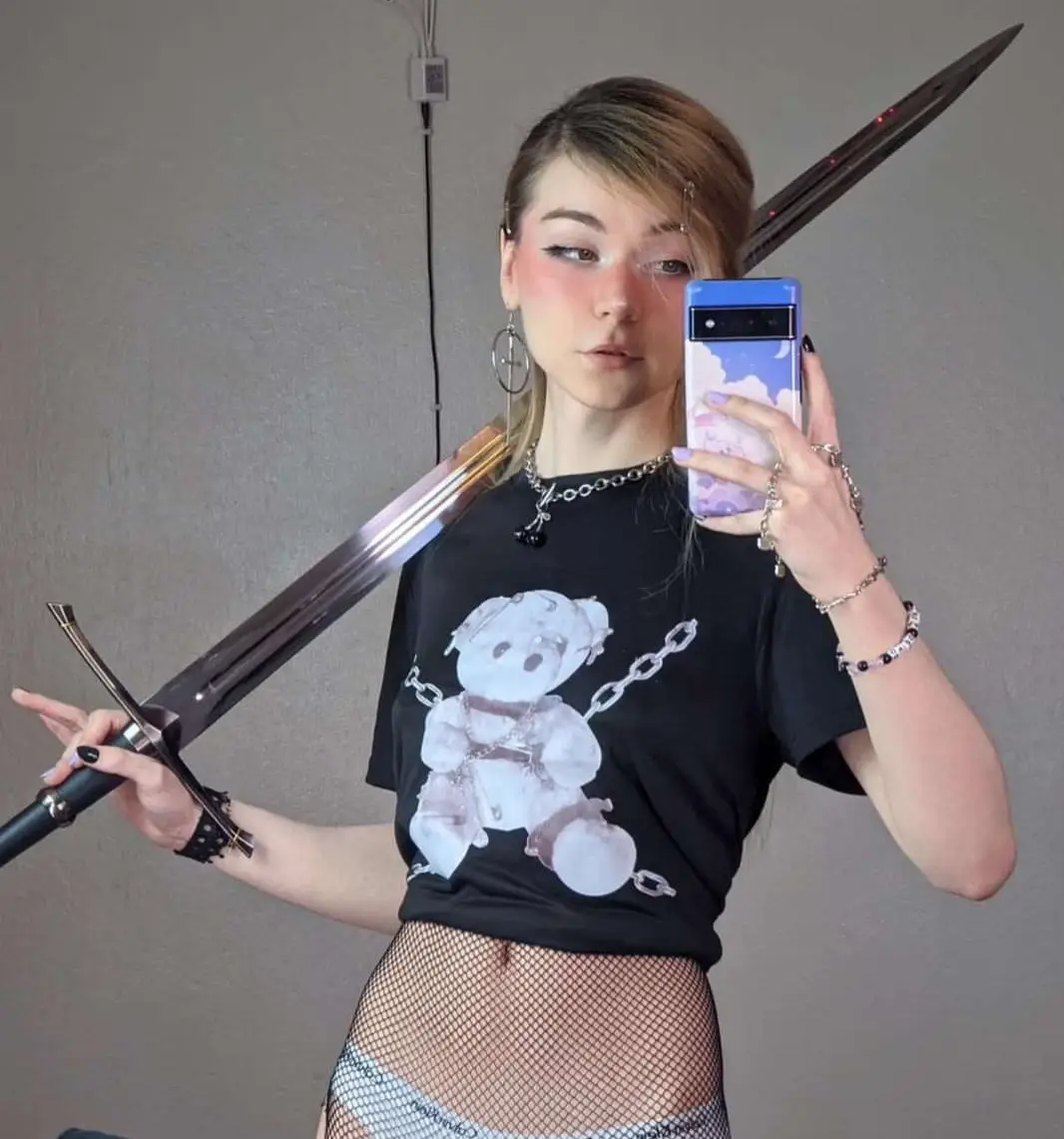 A young woman with blonde hair and heavy makeup takes a mirror selfie holding a large, ornate sword over her shoulder while…