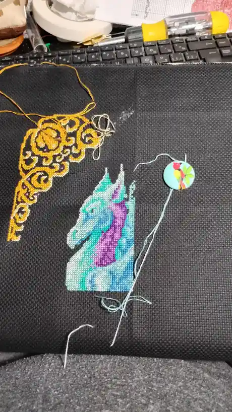 A close-up photo shows a partially completed cross-stitch project on dark fabric, featuring a colorful unicorn head and an…