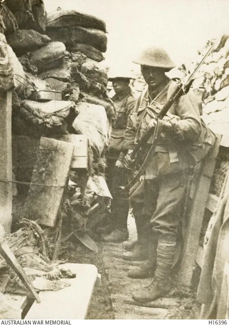 A sepia-toned historical photograph shows two soldiers in World War I-era uniforms and helmets standing in a trench lined…