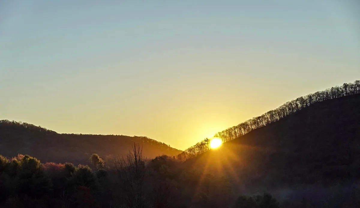 A serene landscape photo shows the sun setting or rising between two forested hills, casting golden light and lens flares…