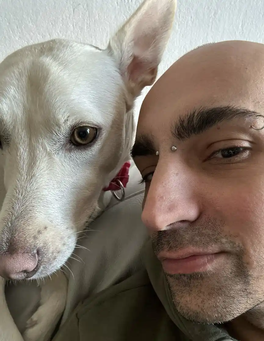 A close-up selfie shows a bald man with facial piercings and a white dog with a pink collar. The man is smiling slightly…