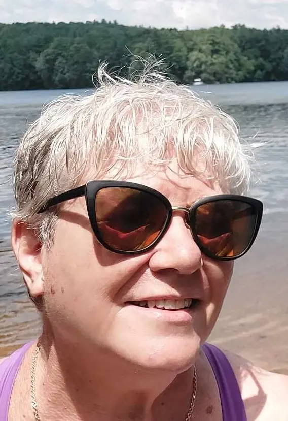 A close-up selfie of a smiling woman with short, wet-looking white hair and large black sunglasses, standing at a lakeshore…