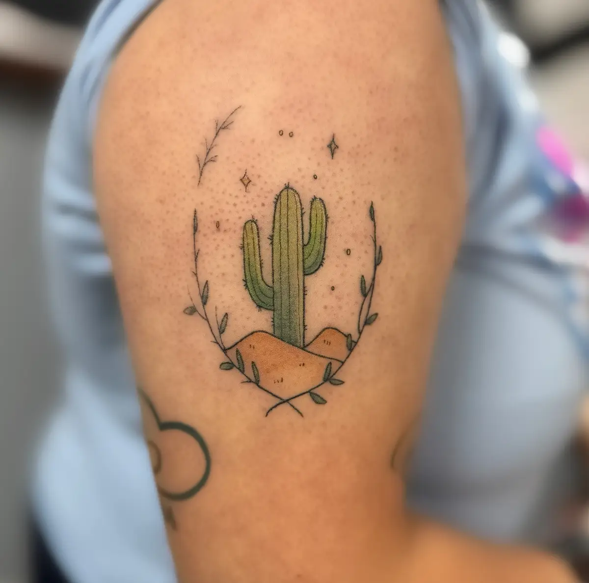A close-up photo shows a tattoo on a person's upper arm featuring a stylized green saguaro cactus with two arms, standing on…
