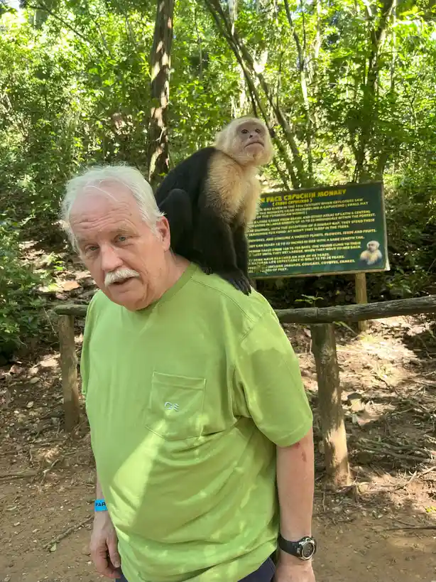 An older man with white hair and a mustache, wearing a green shirt, stands in a forest with a white-faced capuchin monkey…