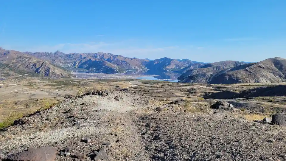 A wide-angle photo shows a barren, rocky foreground leading to a valley with a large, calm lake reflecting distant mountains…