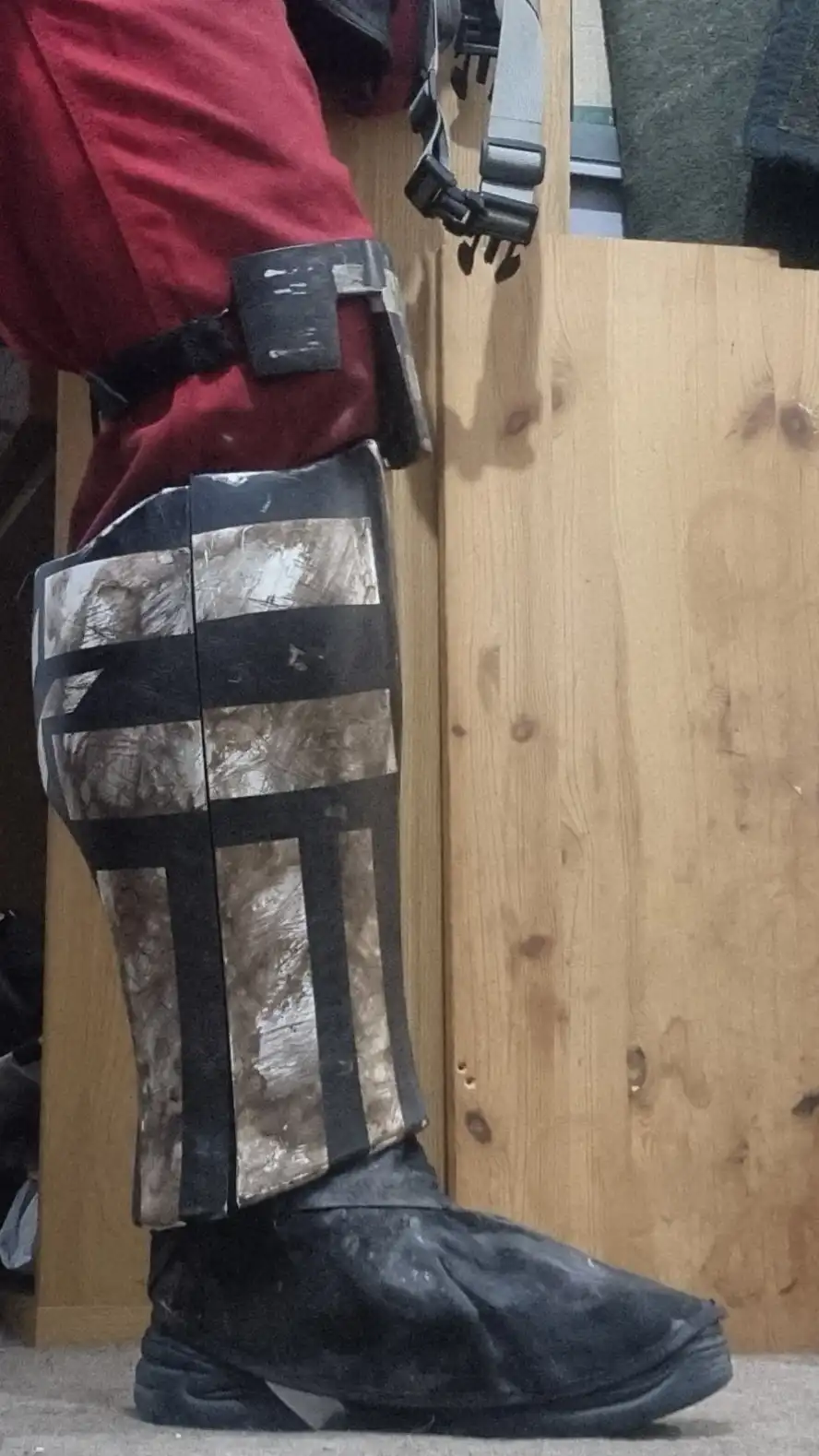 A close-up shot of a person's lower leg wearing a custom-made, heavily worn-looking armored greave over a black boot, paired…