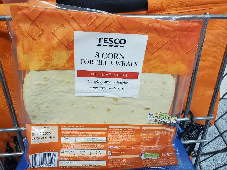 A close-up of a Tesco brand package of 8 corn tortilla wraps, displayed in a supermarket shopping cart. The packaging is…