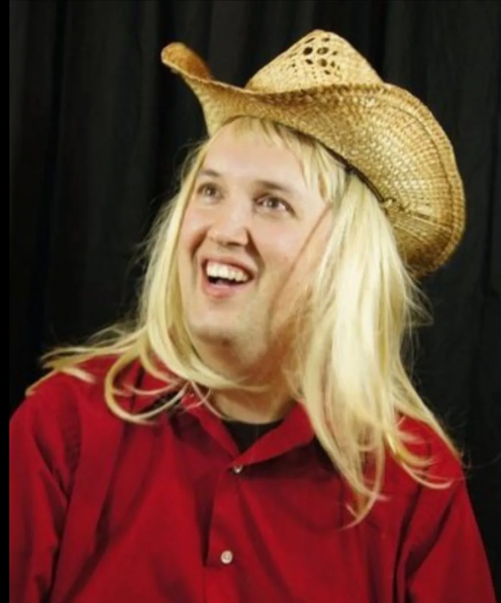 a man with long blonde hair wearing a straw cowboy hat and a red button-up shirt smiles broadly while looking upward against…