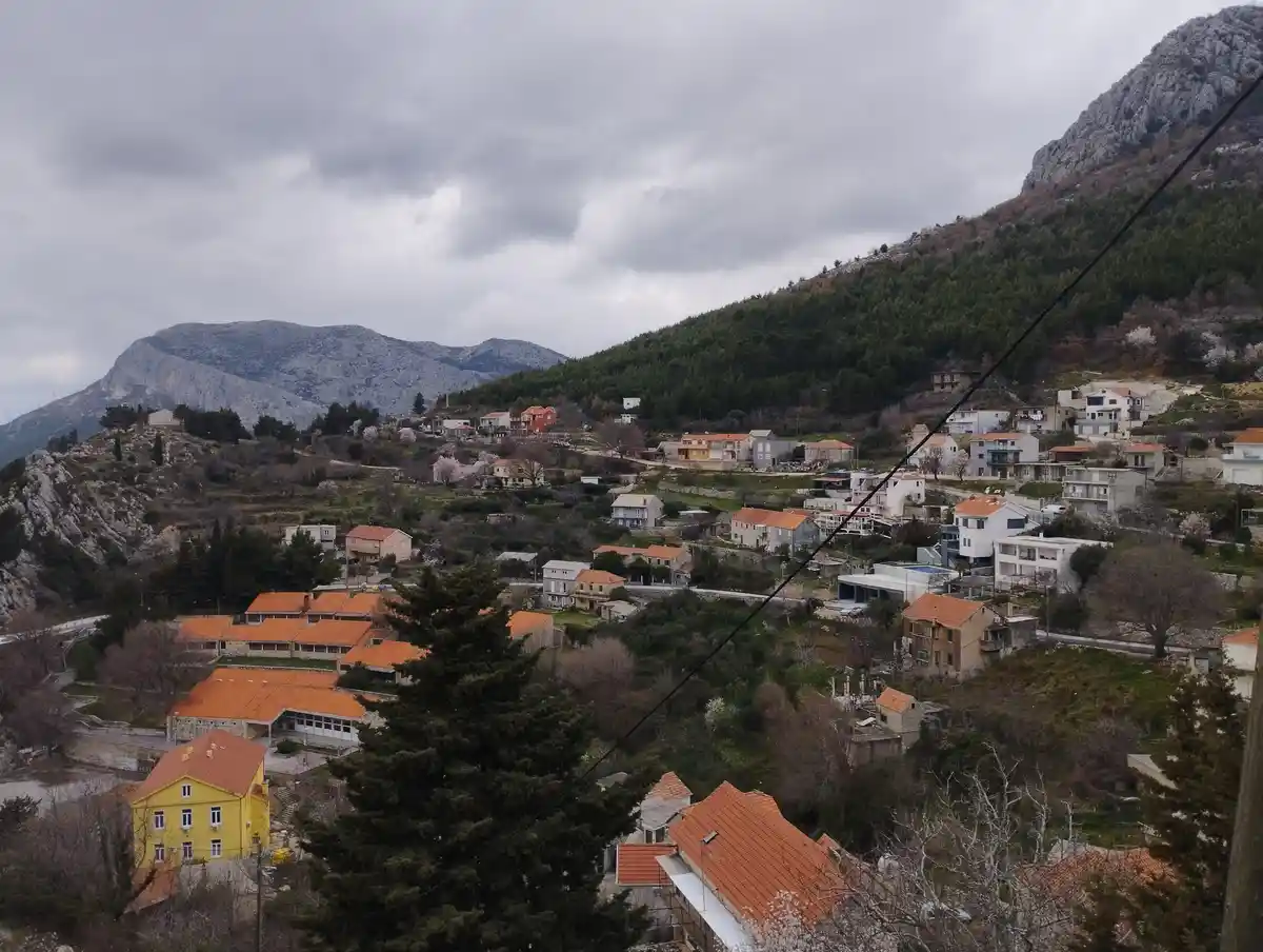 A scenic photo of a village nestled on a hilly, forested landscape under a cloudy sky, featuring houses with orange roofs…
