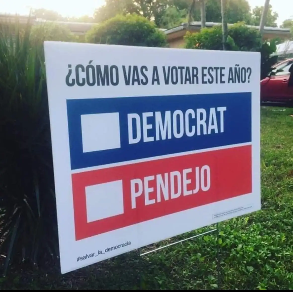 A white yard sign outdoors asks '¿CÓMO VAS A VOTAR ESTE AÑO?' with two options: 'DEMOCRAT' in a blue box and 'PENDEJO' in a…