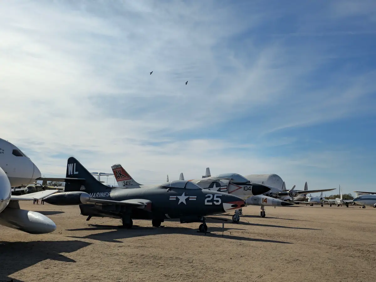 A row of vintage military aircraft sits on a dusty airfield under a partly cloudy sky, with two birds flying in the distance…
