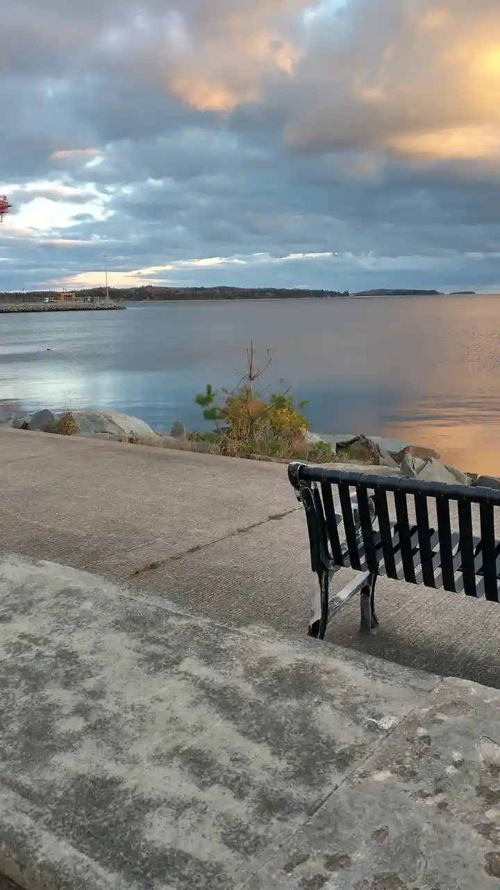A black metal park bench sits on a concrete path overlooking a calm body of water under a cloudy sky with orange sunset…