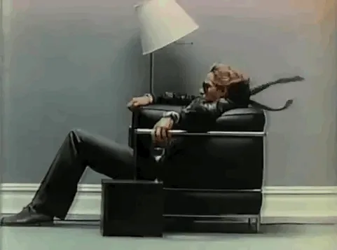 A man in a black suit and sunglasses reclines in a black leather chair, his head tilted back and a long scarf or tie…