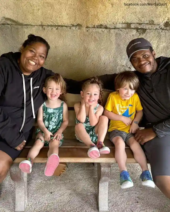 A family photo shows a smiling Black woman and man sitting on either end of a bench with three young children between them…