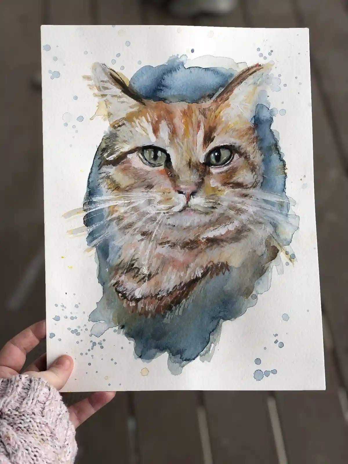 A hand holds up a watercolor painting of a fluffy, orange-and-white cat with green eyes, set against a blue watercolor wash…