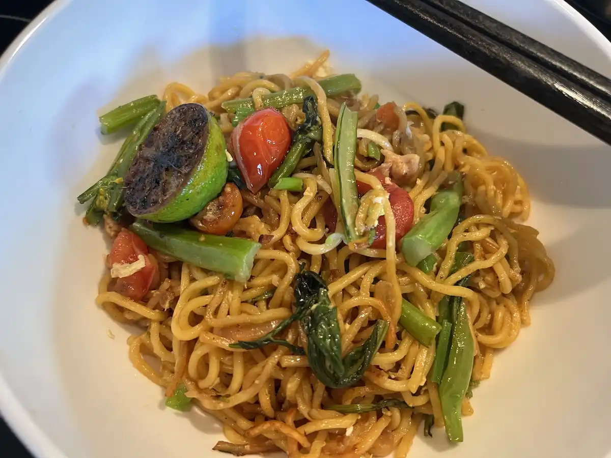 A close-up photo of a white bowl filled with stir-fried noodles mixed with green vegetables, cherry tomatoes, and a charred…