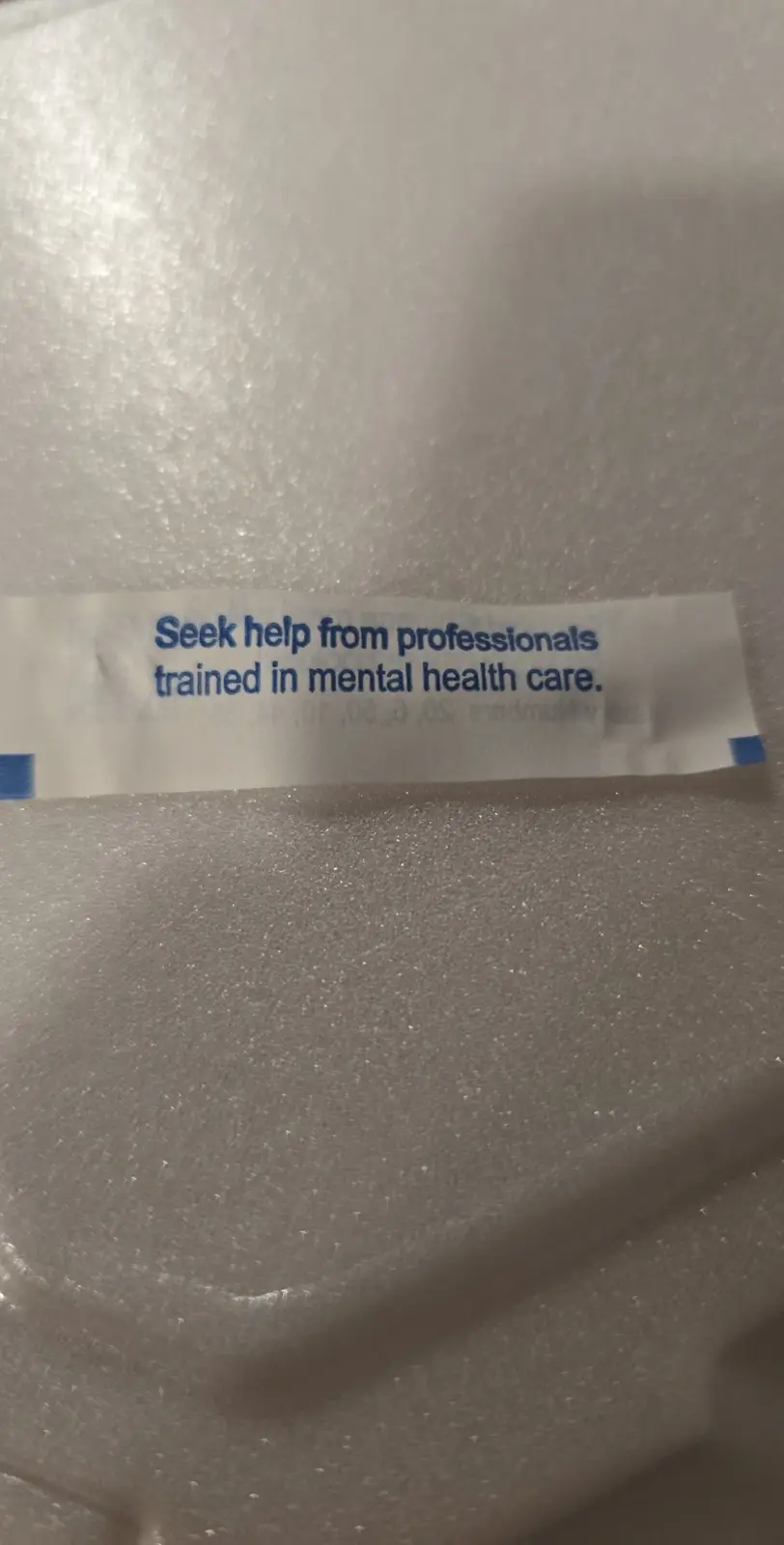 A close-up photo shows a white fortune cookie slip with blue text resting on a textured white foam surface. The text on the…