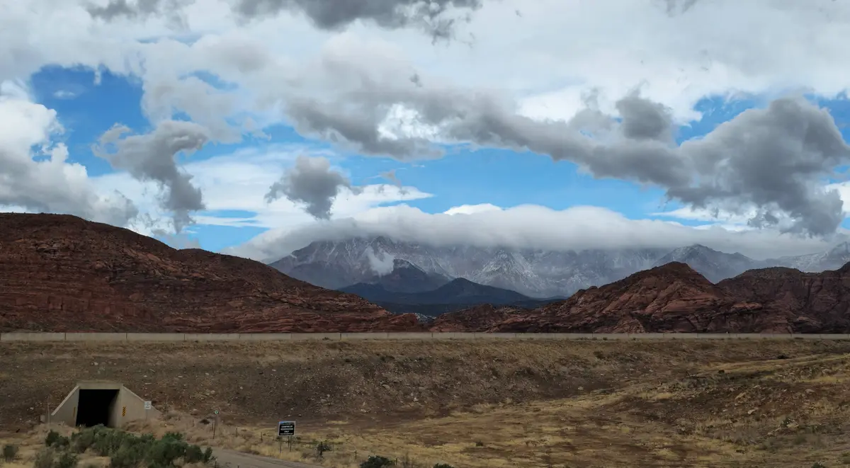 A wide desert landscape with red rock formations and a distant snow-capped mountain range under a partly cloudy sky. In the…