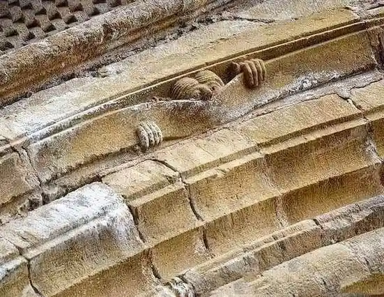 A close-up of ancient stone architecture shows a carved figure with a head and two hands, appearing to be stuck or emerging…