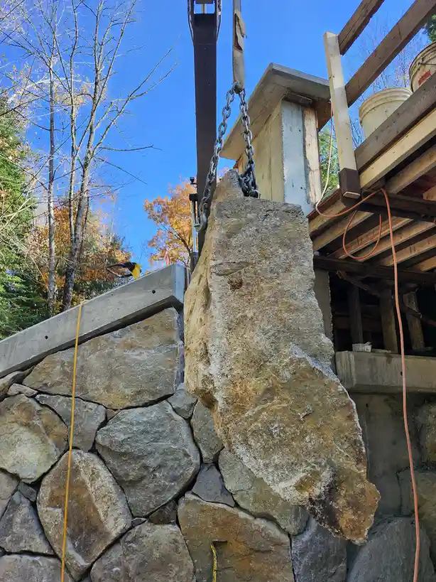 A large, irregularly shaped stone is suspended by chains from a crane, positioned above a partially built stone wall under a…