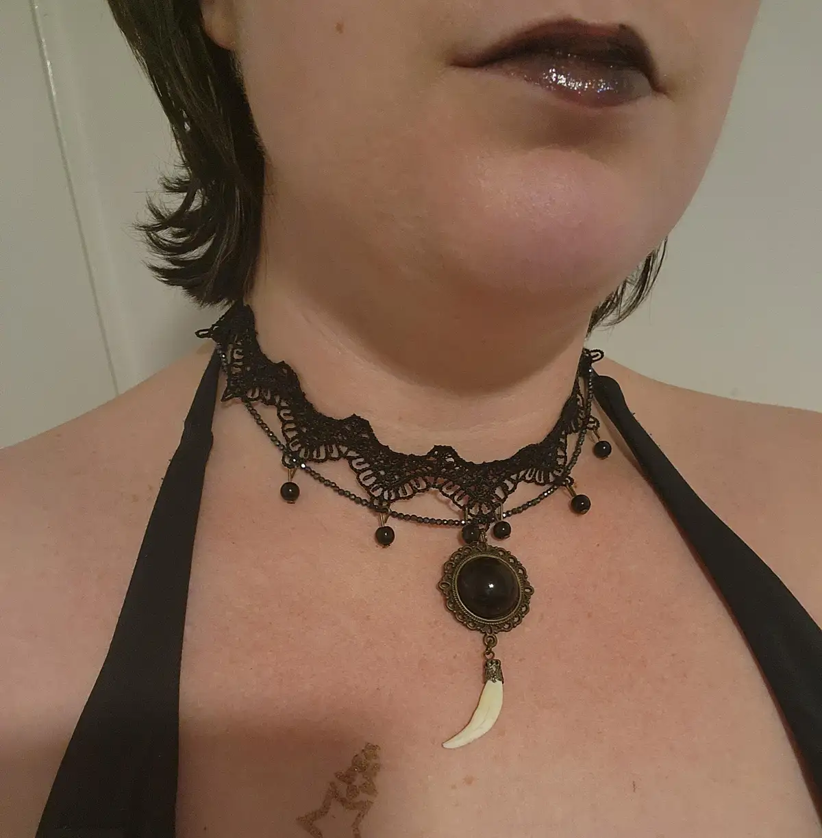 A close-up photo shows a person wearing a black lace choker necklace with black beads and a central dark pendant, from which…