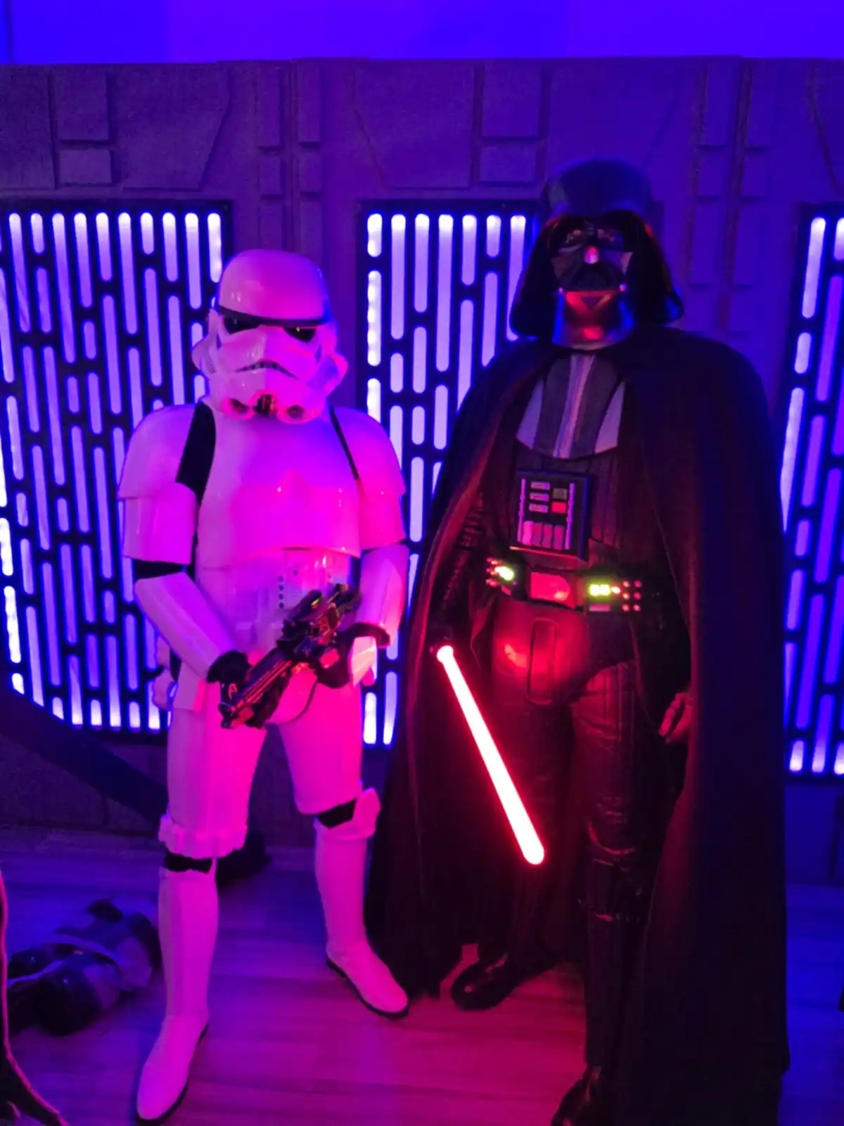 Two people in Star Wars costumes stand side by side under purple lighting: a Stormtrooper holding a blaster and Darth Vader…