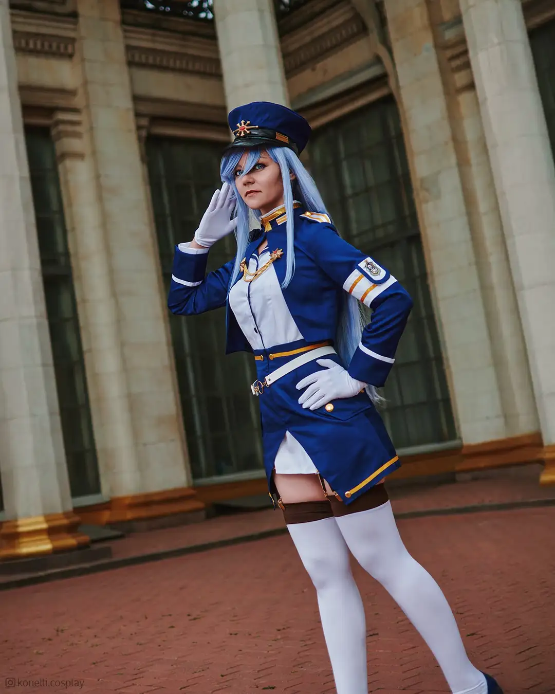 A person with long blue hair is cosplaying as a character in a blue and white military-style uniform with gold trim, white…
