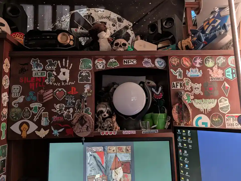 A cluttered wooden desk setup covered in gaming and pop culture stickers, plush toys, and a skull, with a computer monitor…