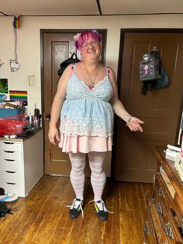 A person with pink hair and glasses stands in a room, wearing a light blue lace top, pink skirt, striped tights, and rainbow…