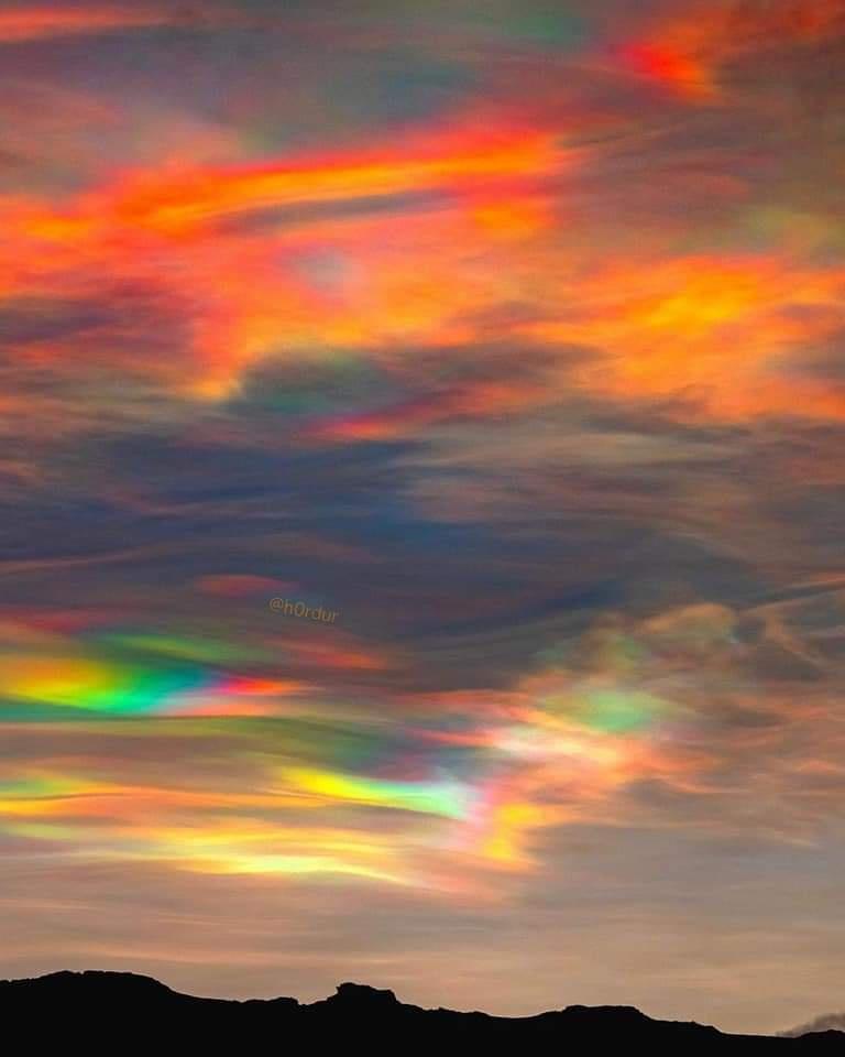 A vibrant, colorful sky with iridescent clouds displaying hues of orange, red, yellow, green, and blue, set above a dark…