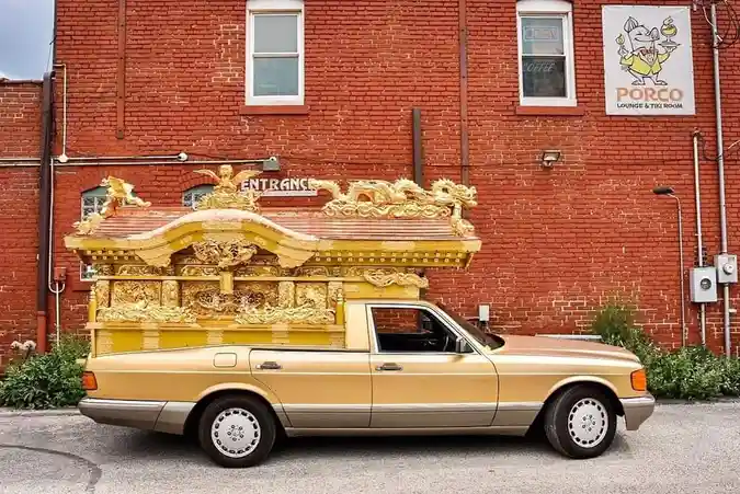 a gold-colored pickup truck has an ornate, gilded Japanese-style coffin mounted on its bed, parked in front of a red brick…
