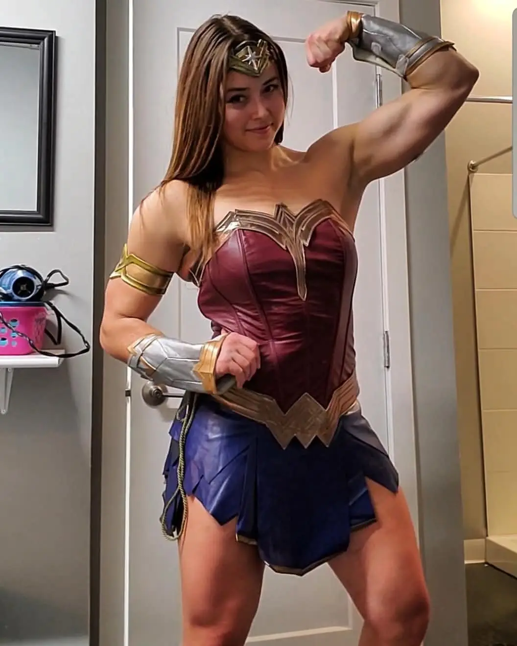 A woman in a detailed Wonder Woman costume, complete with tiara, corset, and gauntlets, poses confidently in a bathroom…
