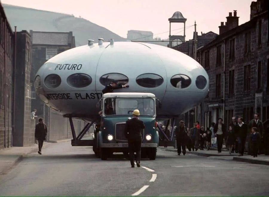 A large, silver, egg-shaped Futuro house, labeled 'FUTURO WATERSIDE PLASTIC', is being transported down a narrow street on a…