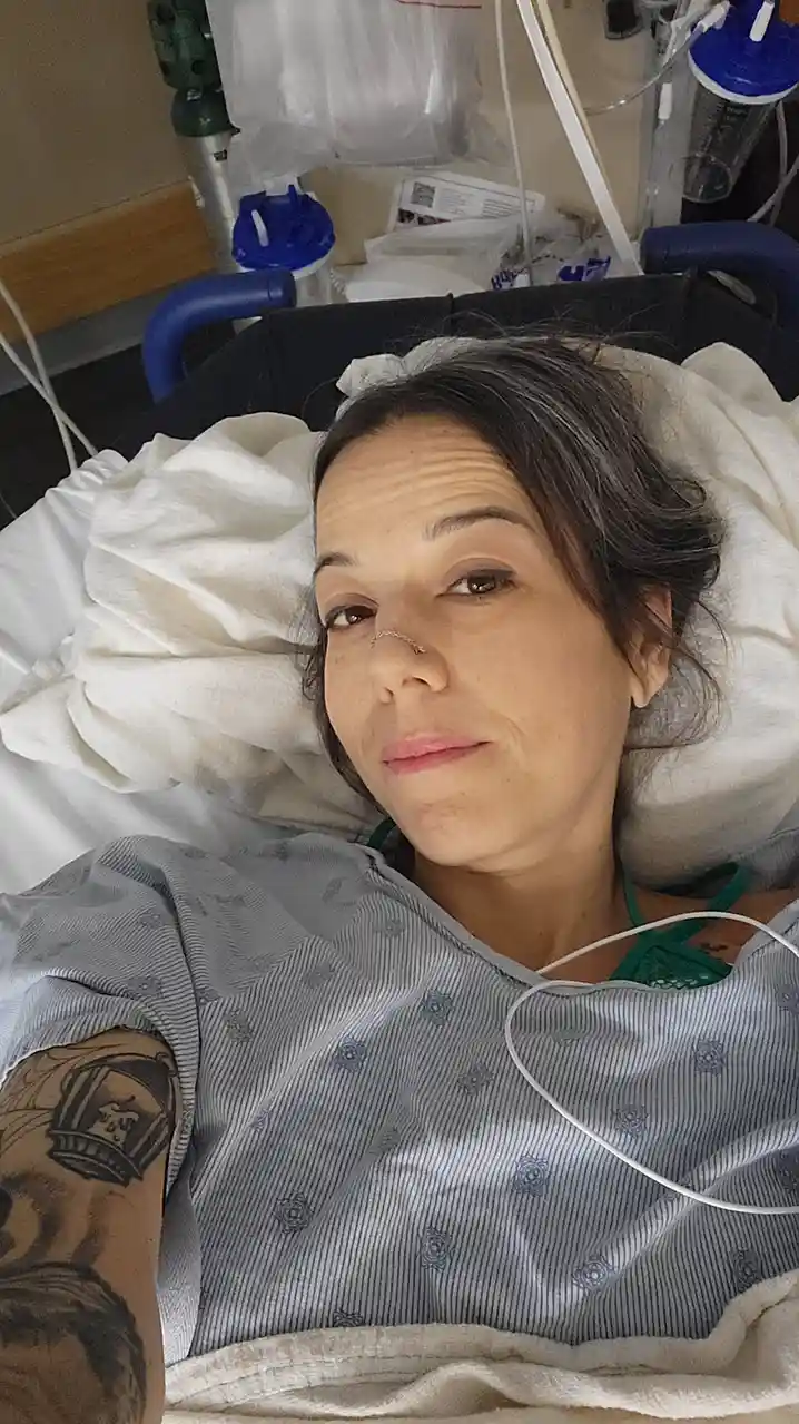 A woman with dark hair and a nose ring lies in a hospital bed wearing a striped gown, surrounded by medical equipment…