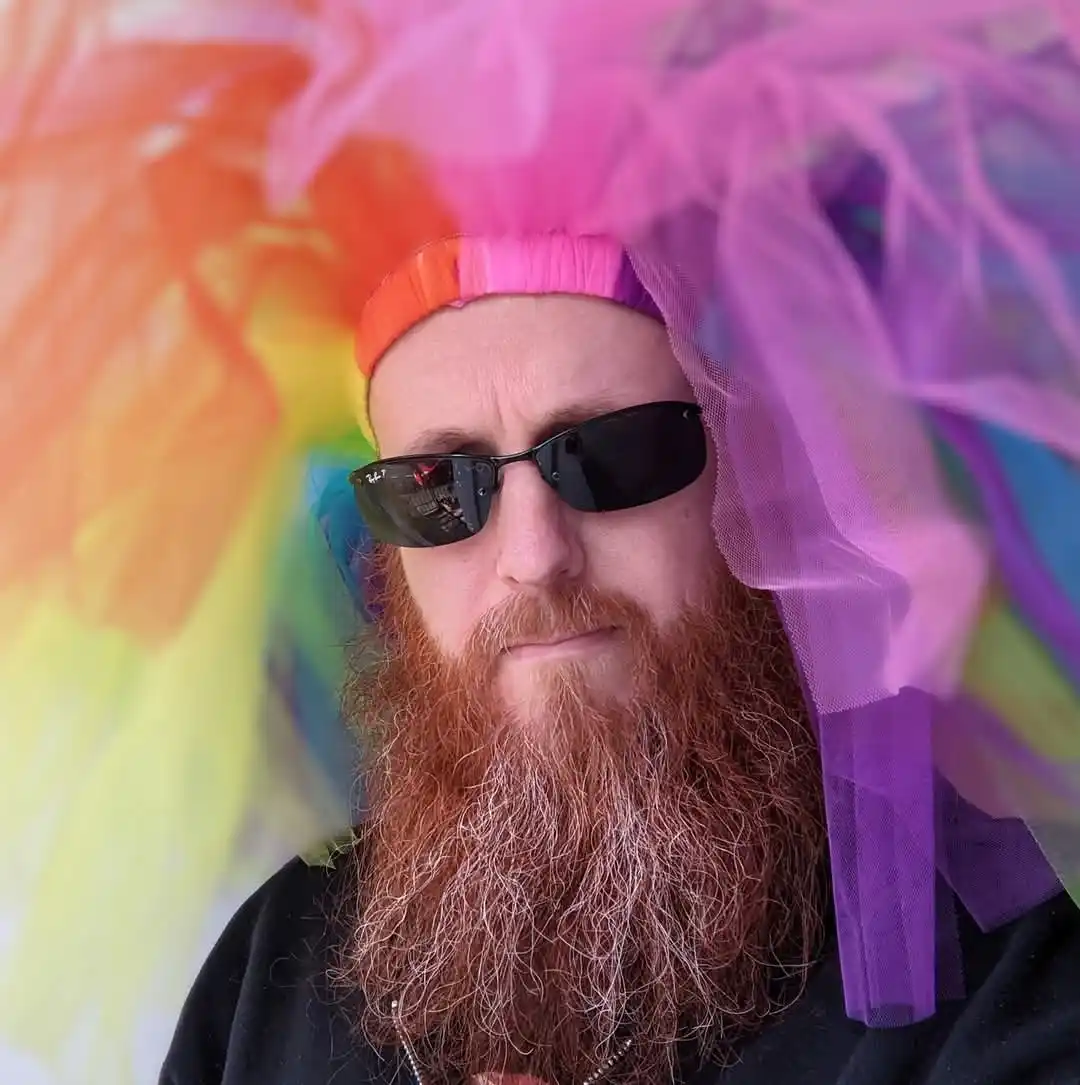 A man with a long red beard and black sunglasses wears a large, colorful rainbow tulle wig. The wig is made of sheer fabric…