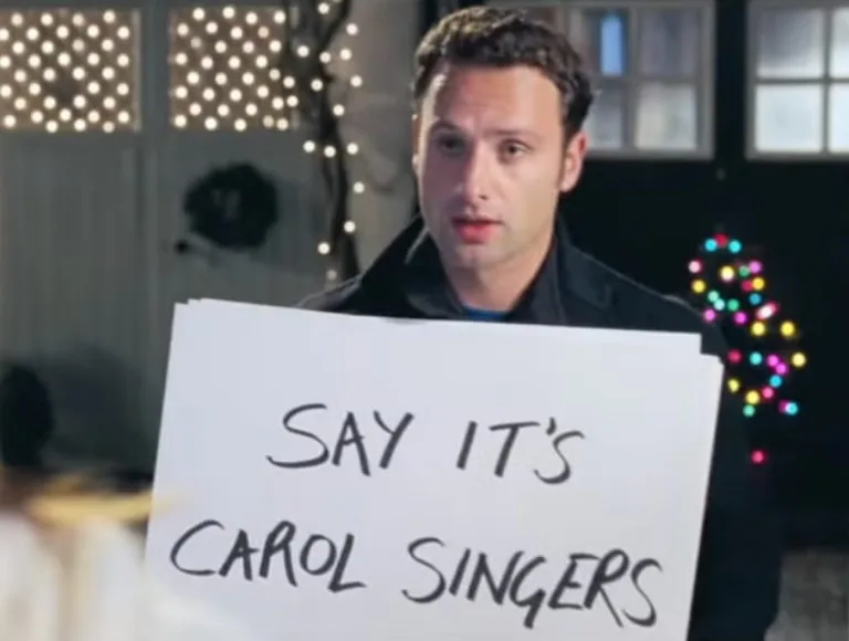A man in a dark jacket holds a white sign with handwritten text that reads 'SAY IT'S CAROL SINGERS'. He looks directly at…