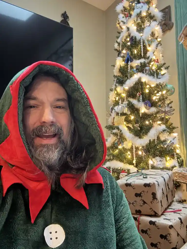 A man with a gray beard wearing a green and red hooded robe with a large white button smiles at the camera. Behind him is a…